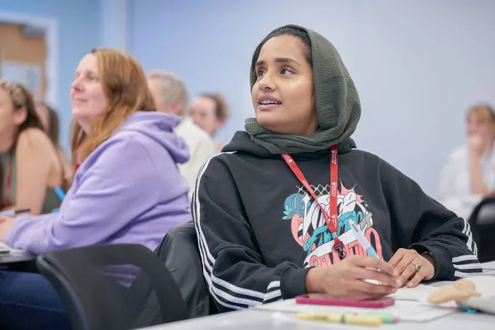 A student wearing a headscarf and black hoodie sits at a desk in a classroom, holding a pen and looking thoughtfully towards the front of the room. Other students are seated nearby, listening and taking notes.