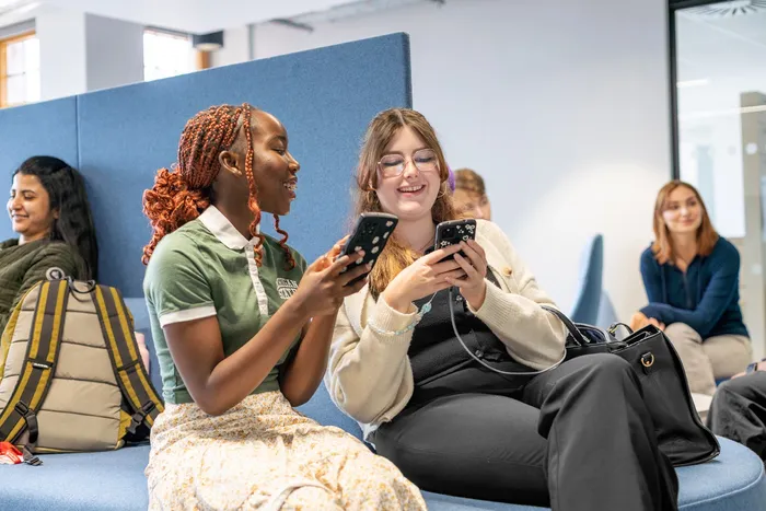 Students in Leeds City Campus Social Space laughing while looking at their phones.