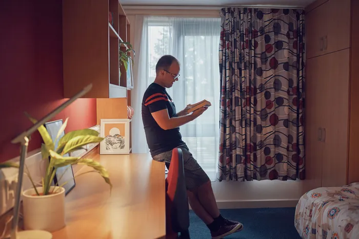 A student leans against a desk in a university accommodation room, reading a book. The room features a long wooden desk with a lamp, plant, and framed artwork, as well as shelving, a wardrobe, and a bed with a patterned duvet. Light filters in through large, patterned curtains covering a window.