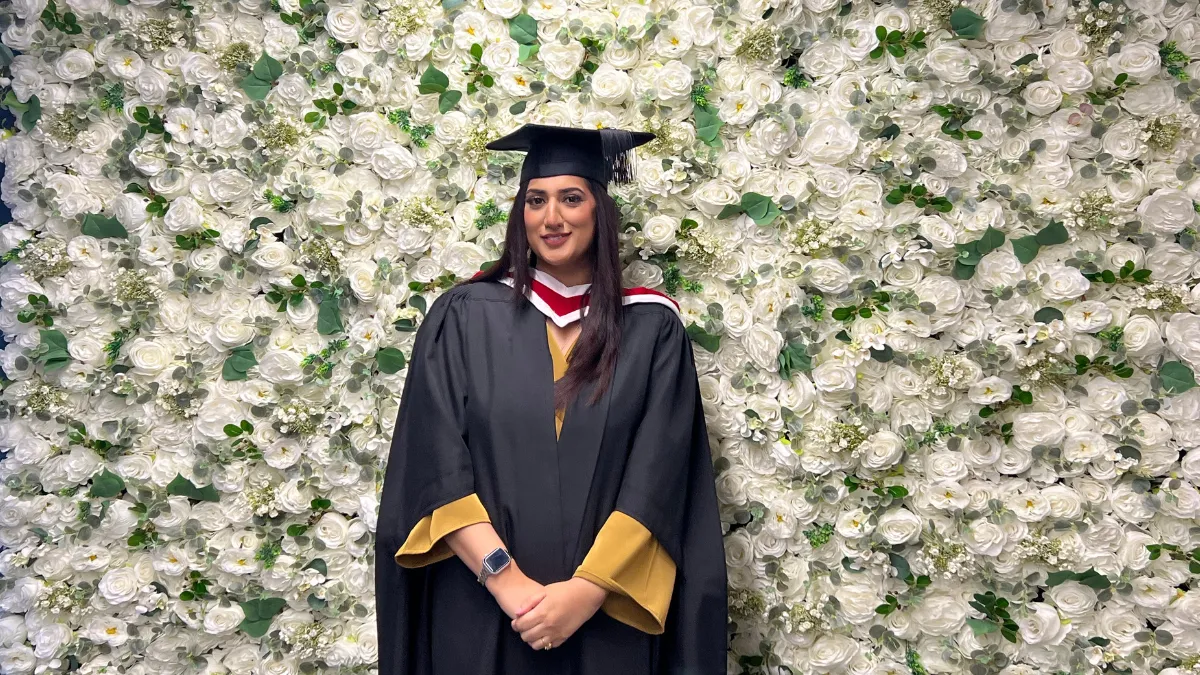 A student in graduation cap and gown smiling against a wall of white flowers.